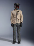 Casaco Alpinestars Borrego Drystar® em Warm Gray, casaco de motociclismo de aventura com quatro bolsos de carga frontais, combinado com calças de condução cinzento escuro, capacete de off-road preto e luvas pretas, com um visual robusto de lona encerada