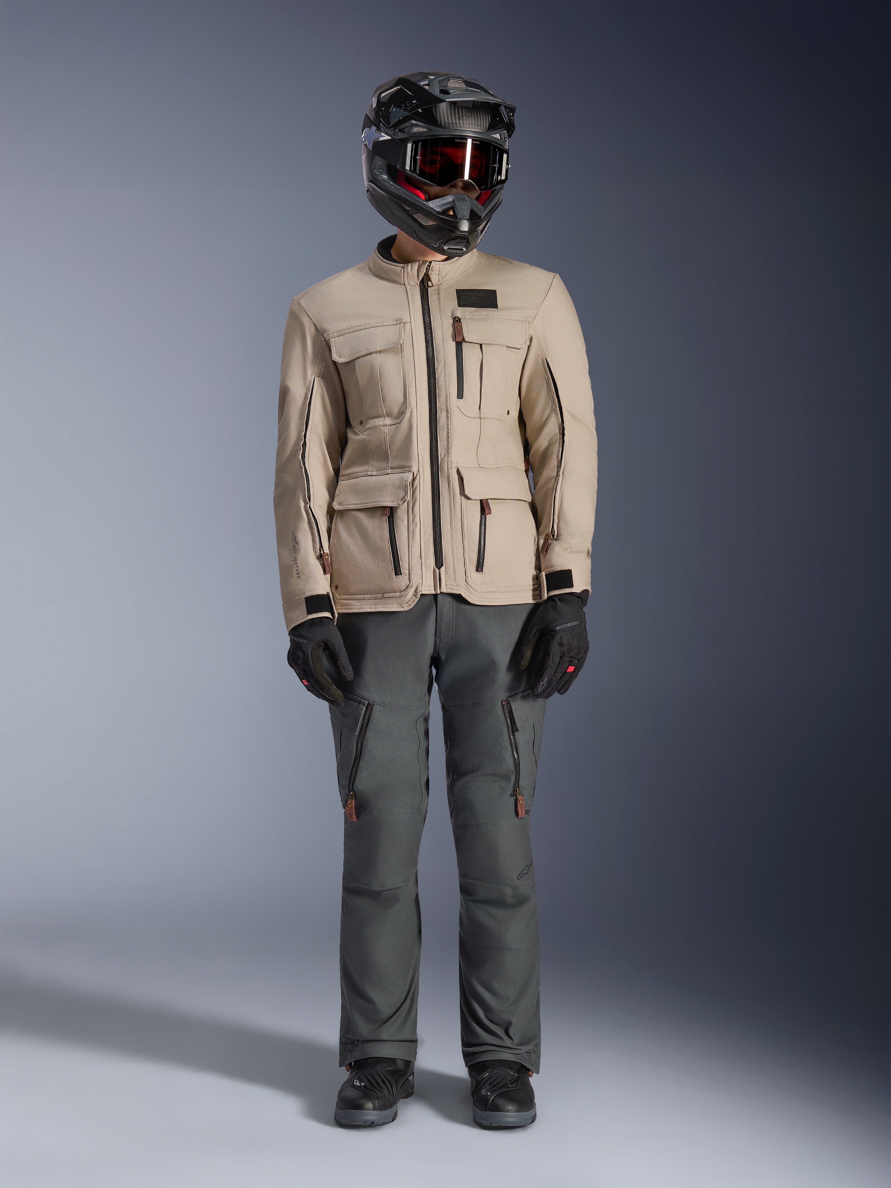 Casaco Alpinestars Borrego Drystar® em Warm Gray, casaco de motociclismo de aventura com quatro bolsos de carga frontais, combinado com calças de condução cinzento escuro, capacete de off-road preto e luvas pretas, com um visual robusto de lona encerada
