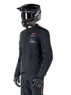 Casaco Alpinestars AMT-8 Stretch Drystar® XF, preto, usado por um motociclista com calças de aventura e luvas a condizer, incluindo capacete de motocross preto e óculos, aberturas de ventilação com fecho no peito e nos braços, marca Alpinestars Motion Technology com logótipo laranja, equipamento premium de adventure-touring