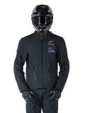 Casaco Alpinestars AMT-8 Stretch Drystar® XF, casaco de motociclismo preto, com logótipo Alpinestars laranja e marca AMT cinzenta, gola alta com fecho frontal e aberturas de ventilação, usado com capacete e luvas pretos, equipamento de proteção para touring de aventura