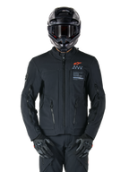 Casaco Alpinestars AMT-8 Stretch Drystar® XF, casaco de motociclismo preto, com logótipo Alpinestars laranja e marca AMT cinzenta, gola alta com fecho frontal e aberturas de ventilação, usado com capacete e luvas pretos, equipamento de proteção para touring de aventura