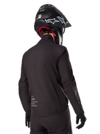 Alpinestars AMT-8 Stretch Drystar® XF Liner, forro de casaco impermeável preto, vista traseira num motociclista com capacete e luvas pretos, com o logótipo Alpinestars Motion Technology na zona lombar, design minimalista e funcional para condução de aventura