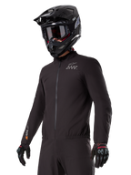 Piloto a usar o forro Alpinestars AMT-8 Stretch Drystar® XF, casaco elástico preto à prova de vento e impermeável, com o logótipo AMT no peito e a marca Drystar XF na manga, combinado com um capacete de aventura preto e luvas de proteção.