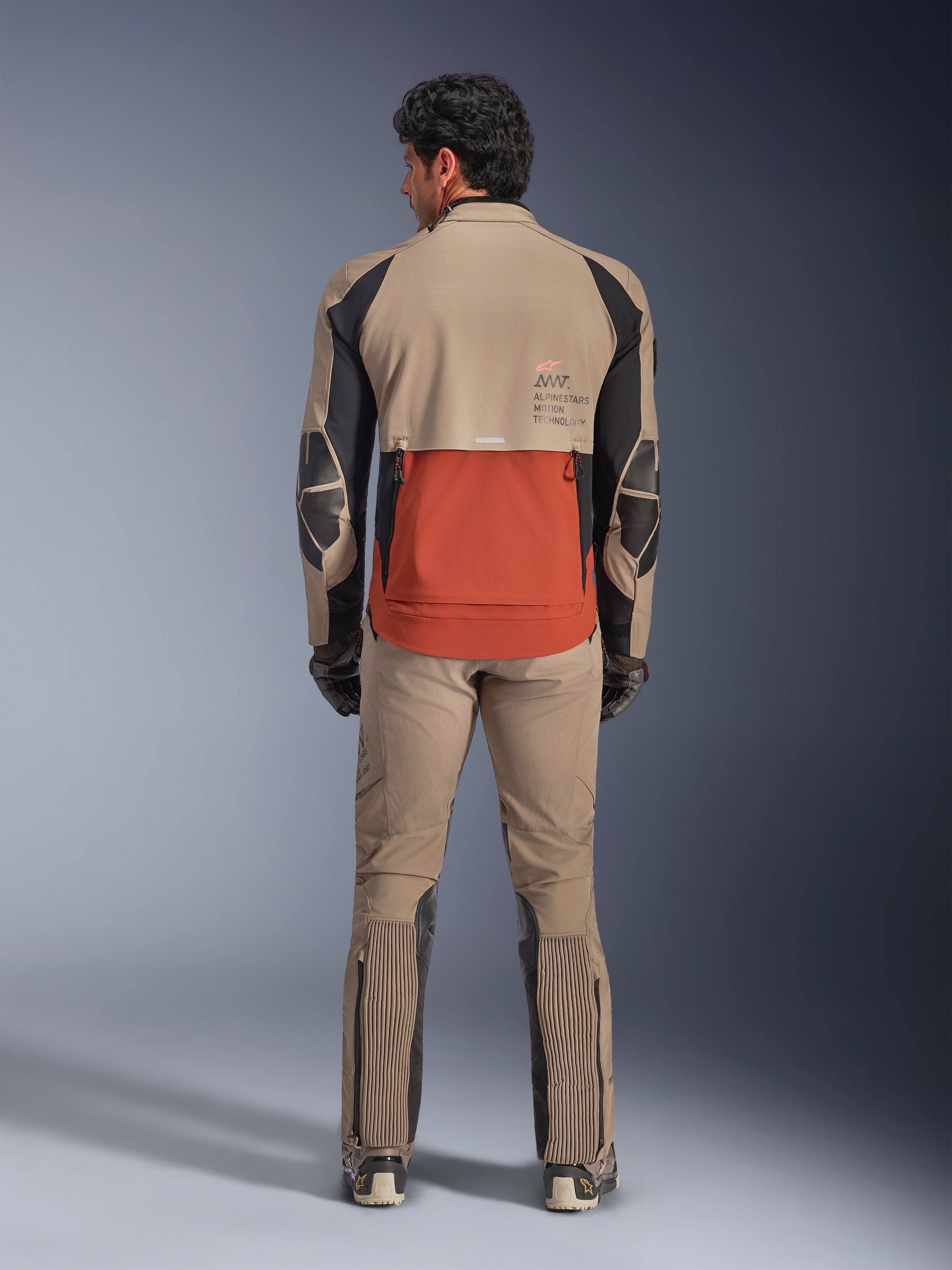 Alpinestars AMT-8 Stretch Drystar® XF Jacket, Walnut Rust Black, vista traseira de um motociclista com calças de aventura e luvas a condizer, apresentando um painel traseiro proeminente cor de ferrugem, secções superiores nogueira e detalhes elásticos pretos, concebido para touring de longa distância