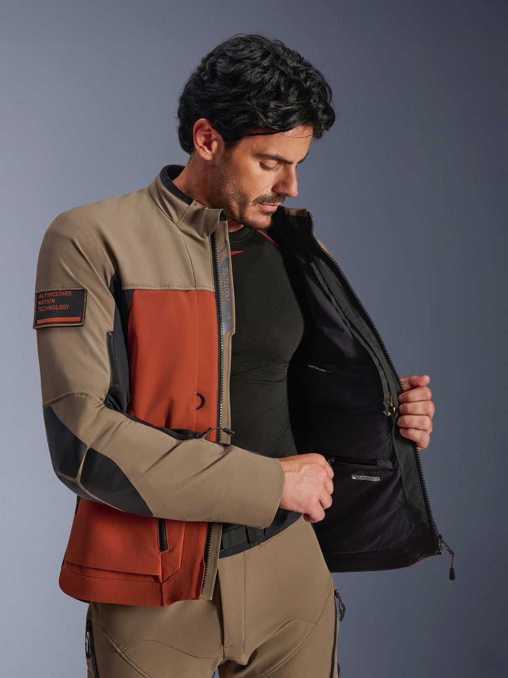 Casaco Alpinestars AMT-8 Stretch Drystar® XF, Walnut Rust Black, homem a usar o casaco aberto para mostrar o compartimento interior impermeável e o forro técnico, com o emblema 'Alpinestars Motion Technology' no braço, equipamento de motociclismo de aventura.
