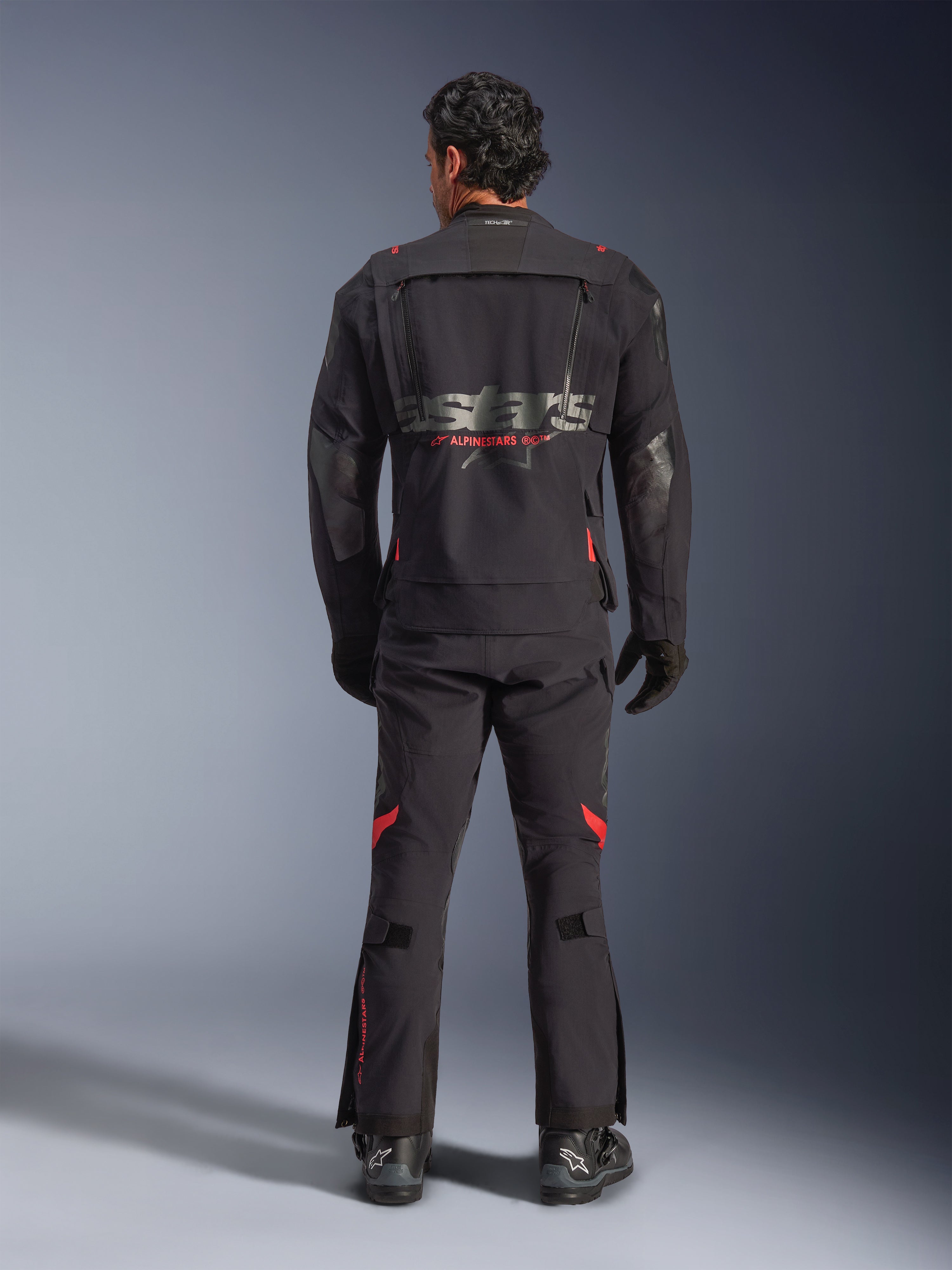 Vista traseira de um motociclista com o Casaco Alpinestars Halo Pro Drystar® XF Laminated em Black Dark Shadow, com um grande logótipo astars cinzento e marca Alpinestars a vermelho, painéis de ventilação modulares nas costas, calças de aventura a condizer e botas pretas.