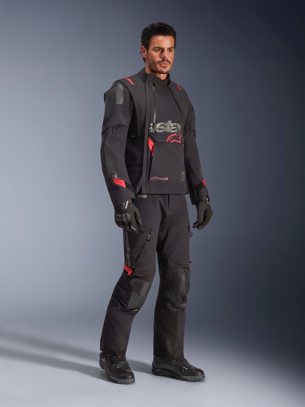 Motociclista com o Casaco Laminado Alpinestars Halo Pro Drystar® XF em Preto Dark Shadow, equipamento de aventura completo com calças, luvas e botas a condizer, apresentando painéis frontais assimétricos modulares e detalhes da marca em vermelho.