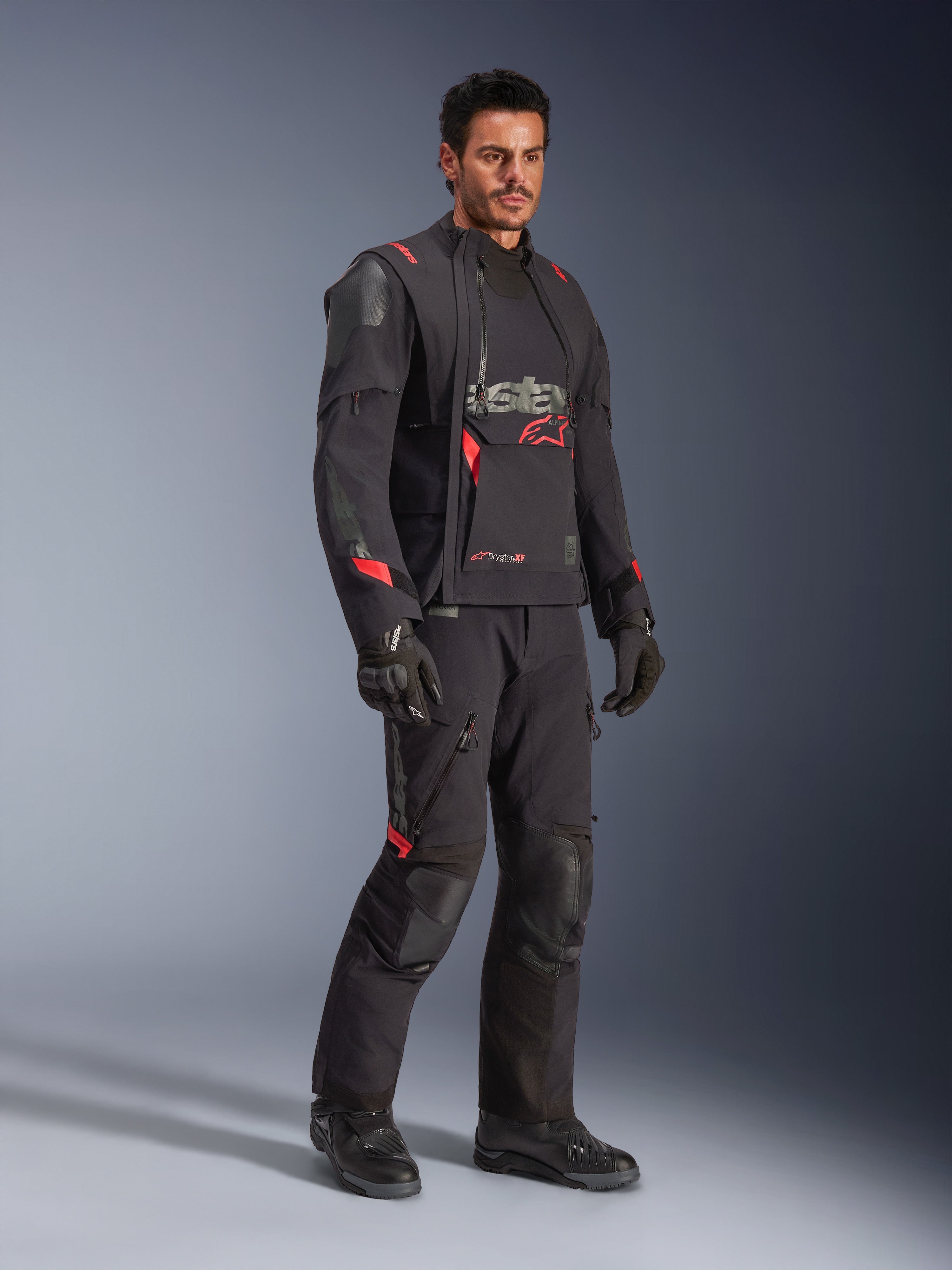 Motociclista com o Casaco Laminado Alpinestars Halo Pro Drystar® XF em Preto Dark Shadow, equipamento de aventura completo com calças, luvas e botas a condizer, apresentando painéis frontais assimétricos modulares e detalhes da marca em vermelho.
