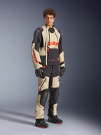 Modelo masculino a usar o Casaco Alpinestars Halo Pro Drystar® XF Laminated em Mastic Black, com painéis bege e pretos e detalhes em vermelho, combinado com calças de aventura a condizer e luvas pretas, concebido para motociclismo de aventura em todo-o-terreno.