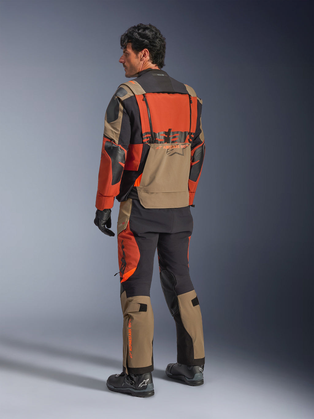 Casaco Laminado Alpinestars Halo Pro Drystar® XF, casaco de motociclismo de aventura em Walnut Rust Black, vista traseira usado com calças a condizer e botas pretas, com material elástico técnico 3L, painéis de ventilação modulares e logótipo cor de laranja na parte traseira