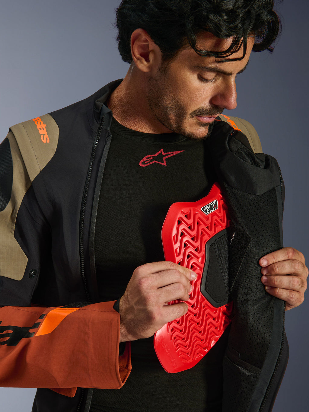 Alpinestars Halo Pro Drystar® XF Laminated Jacket em Walnut Rust Black, fotografia de estilo de vida de um modelo masculino a abrir o fecho do casaco de aventura para revelar uma proteção de peito vermelha, com marca em laranja no ombro e uma camada de base preta com logótipo vermelho.