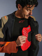 Alpinestars Halo Pro Drystar® XF Laminated Jacket em Walnut Rust Black, fotografia de estilo de vida de um modelo masculino a abrir o fecho do casaco de aventura para revelar uma proteção de peito vermelha, com marca em laranja no ombro e uma camada de base preta com logótipo vermelho.