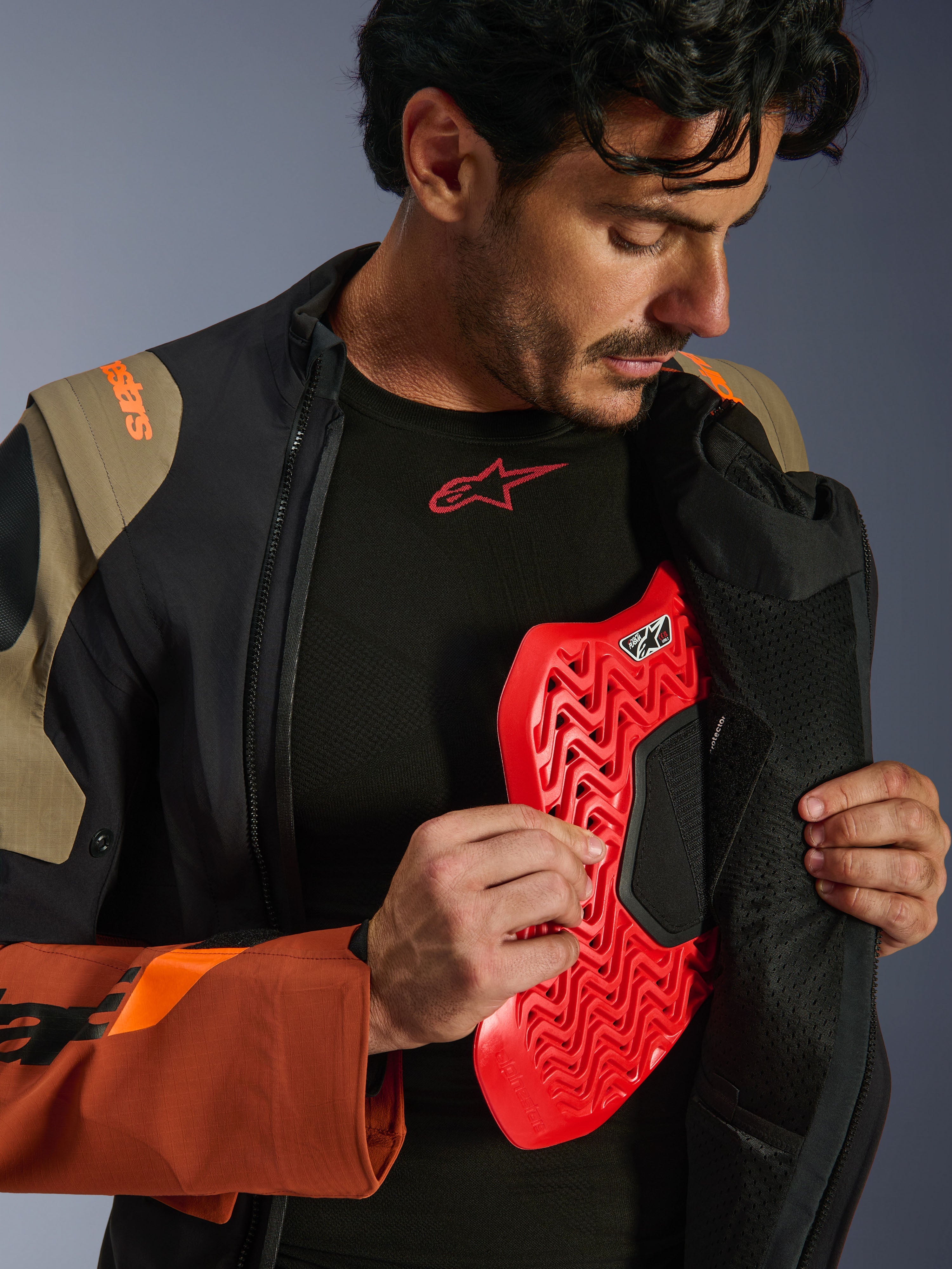 Alpinestars Halo Pro Drystar® XF Laminated Jacket em Walnut Rust Black, fotografia de estilo de vida de um modelo masculino a abrir o fecho do casaco de aventura para revelar uma proteção de peito vermelha, com marca em laranja no ombro e uma camada de base preta com logótipo vermelho.