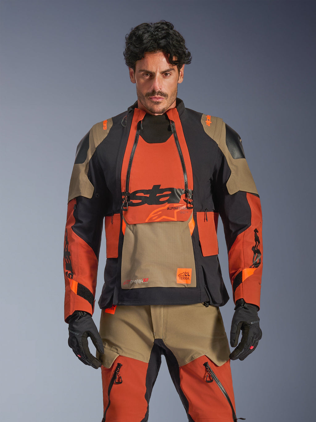 Casaco Alpinestars Halo Pro Drystar XF Laminado, casaco de motociclismo para turismo de aventura, castanho Walnut Rust e preto com pormenores bege, usado por um modelo com equipamento a condizer, vista frontal, com construção laminada impermeável e painéis de ventilação modular no peito, equipamento de condução premium para todo-o-terreno