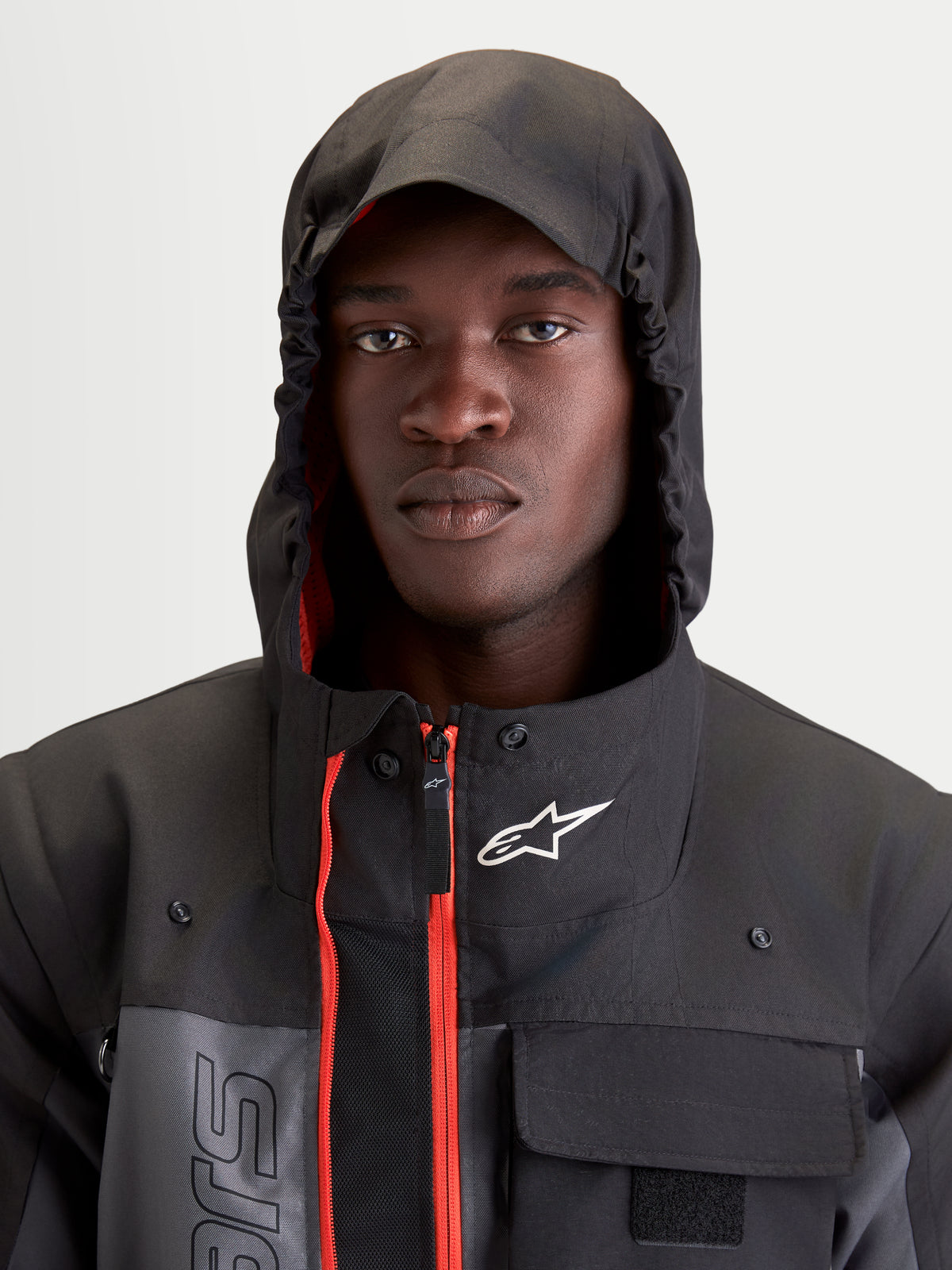 Alpinestars Morfex Parka Waterproof Jacket, casaco de moto urbano e contemporâneo para homem em cinzento e preto, retrato em plano aproximado de um modelo a vestir o casaco com capuz integrado colocado e com detalhes interiores vermelhos visíveis