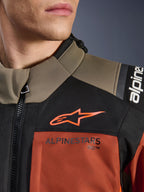 Casaco Alpinestars Andes V4 Drystar® em Walnut Rust Black, grande plano da parte superior do peito e ombro, com logótipo A-star cor de laranja no painel preto e marca Alpinestars na secção cor de ferrugem, detalhe de equipamento de motociclismo de aventura.