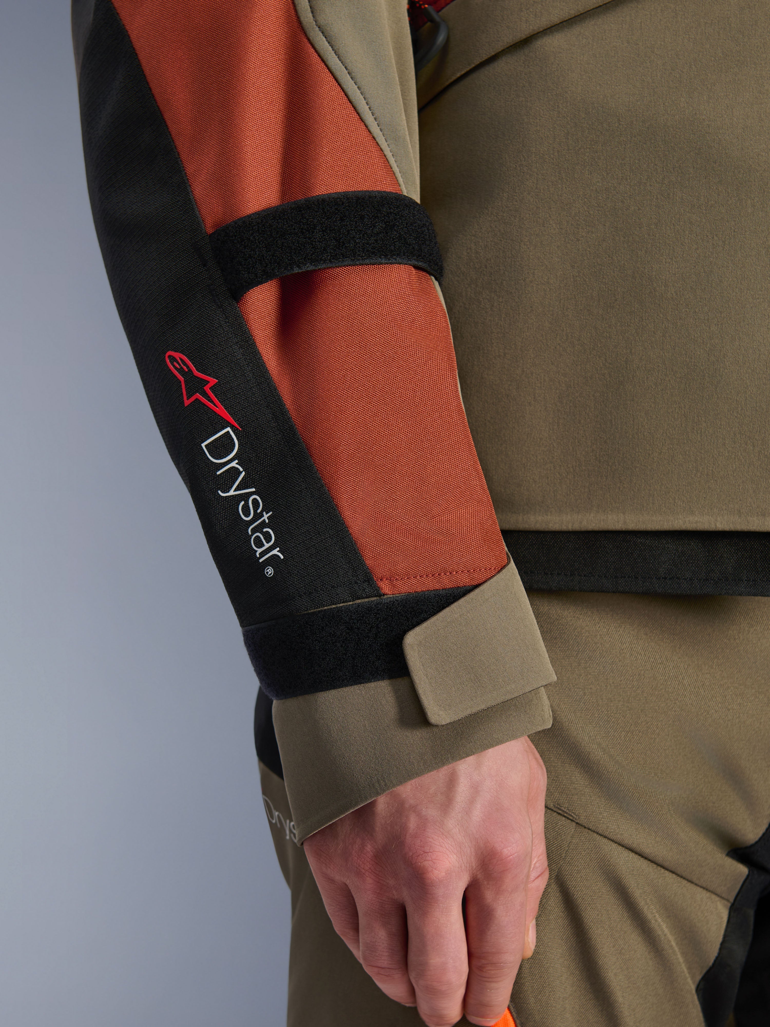 Casaco Alpinestars Andes V4 Drystar®, casaco de motociclismo, Walnut Rust Black, vista de pormenor da manga com a marca Drystar® e o logótipo da estrela vermelha, com tiras de velcro ajustáveis no antebraço e no pulso, design têxtil durável para adventure touring