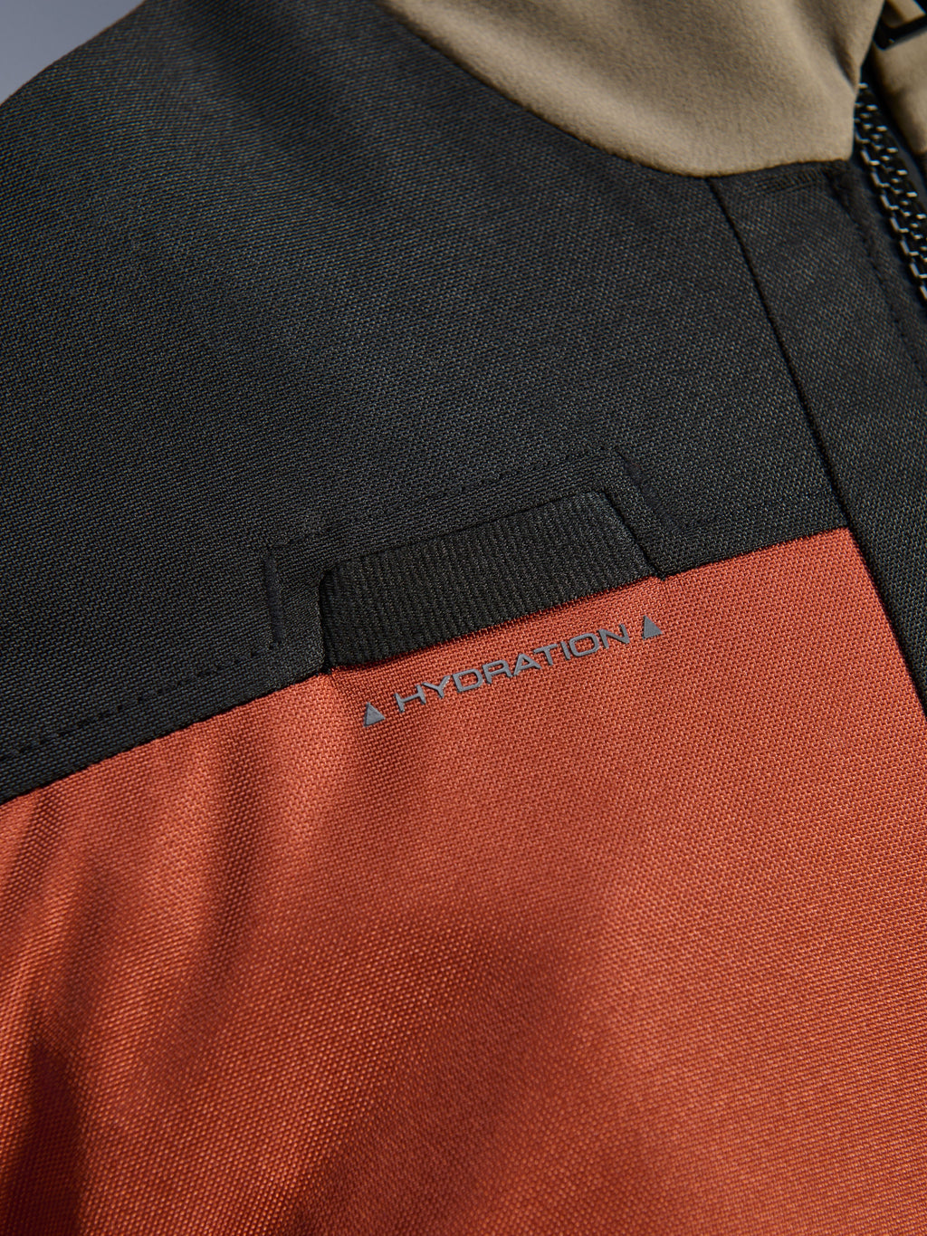 Pormenor em grande plano do Casaco Alpinestars Andes V4 Drystar® em Walnut Rust Black, destacando o laço integrado para mangueira de hidratação no peito e a construção têxtil em ripstop durável.