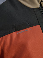 Pormenor em grande plano do Casaco Alpinestars Andes V4 Drystar® em Walnut Rust Black, destacando o laço integrado para mangueira de hidratação no peito e a construção têxtil em ripstop durável.