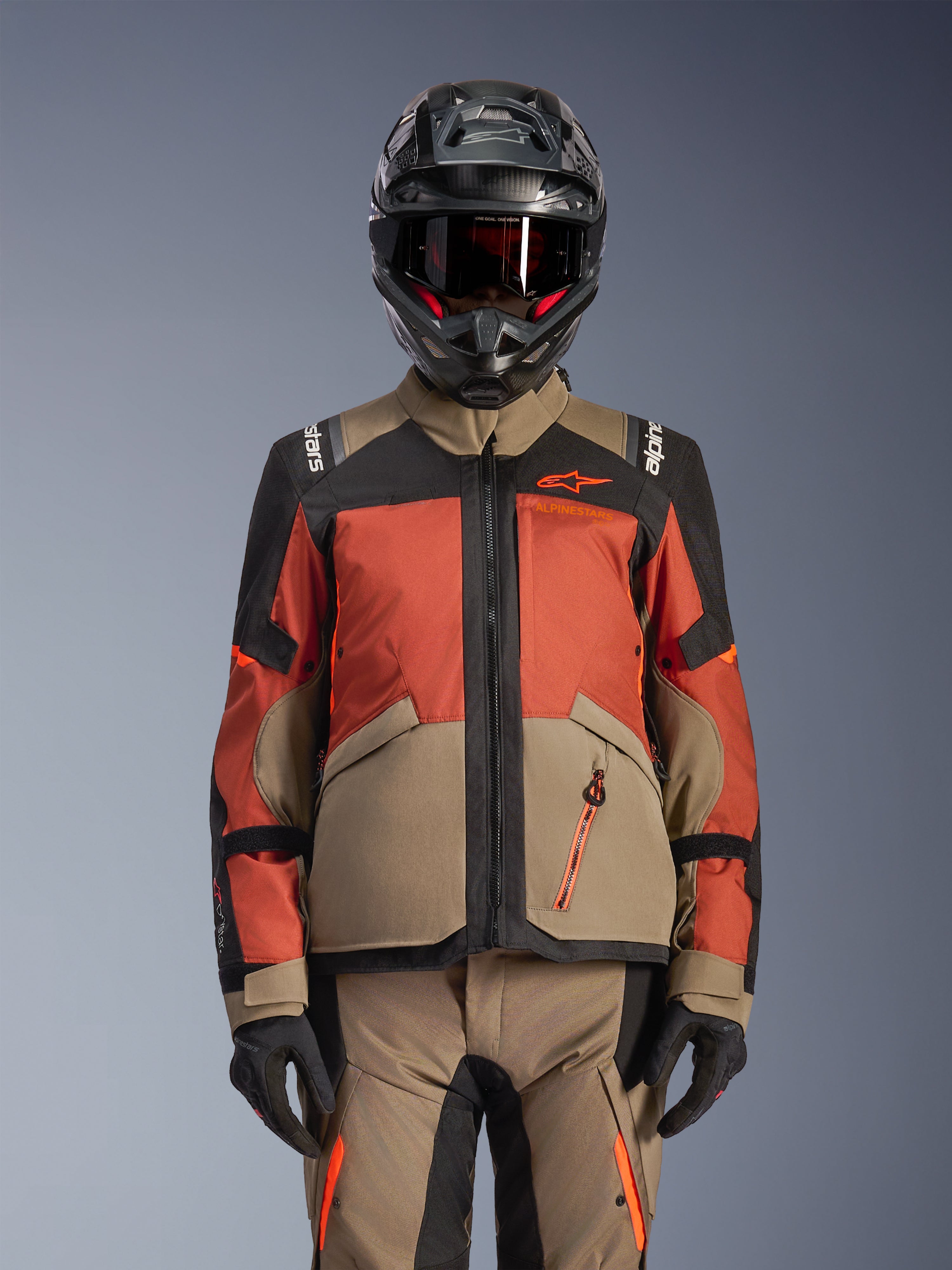 Casaco Alpinestars Andes V4 Drystar®, Casaco de Motociclismo de Aventura, Walnut Rust Black, usado com capacete e luvas a condizer, com um design técnico multi-painel em bege, laranja avermelhado e preto, logótipo laranja visível e fechos de correr impermeáveis.