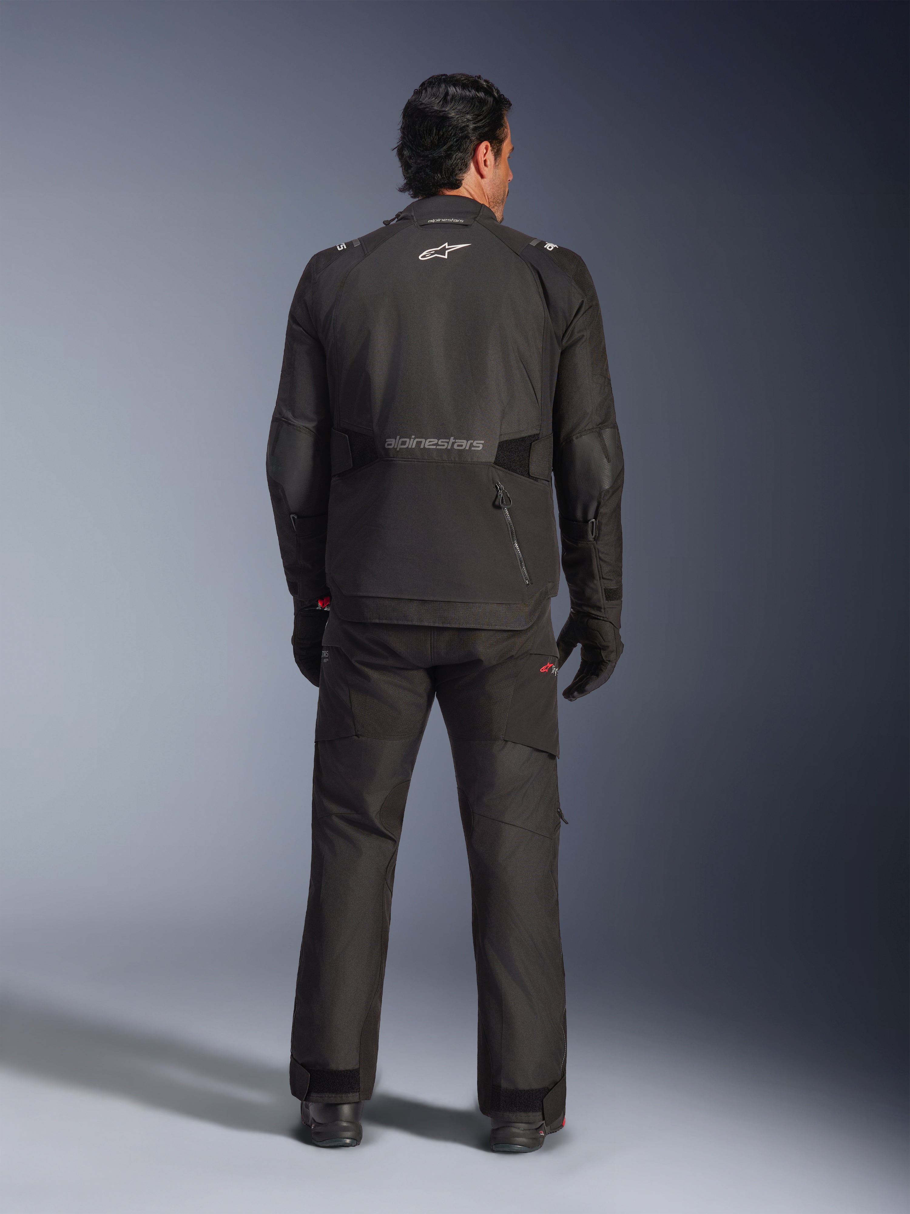 Vista traseira de um motociclista com o Casaco Alpinestars Andes V4 Drystar® em preto, combinado com calças e luvas de motociclismo a condizer, com o logótipo e a inscrição Alpinestars nas costas, equipamento técnico de aventura para todas as condições meteorológicas