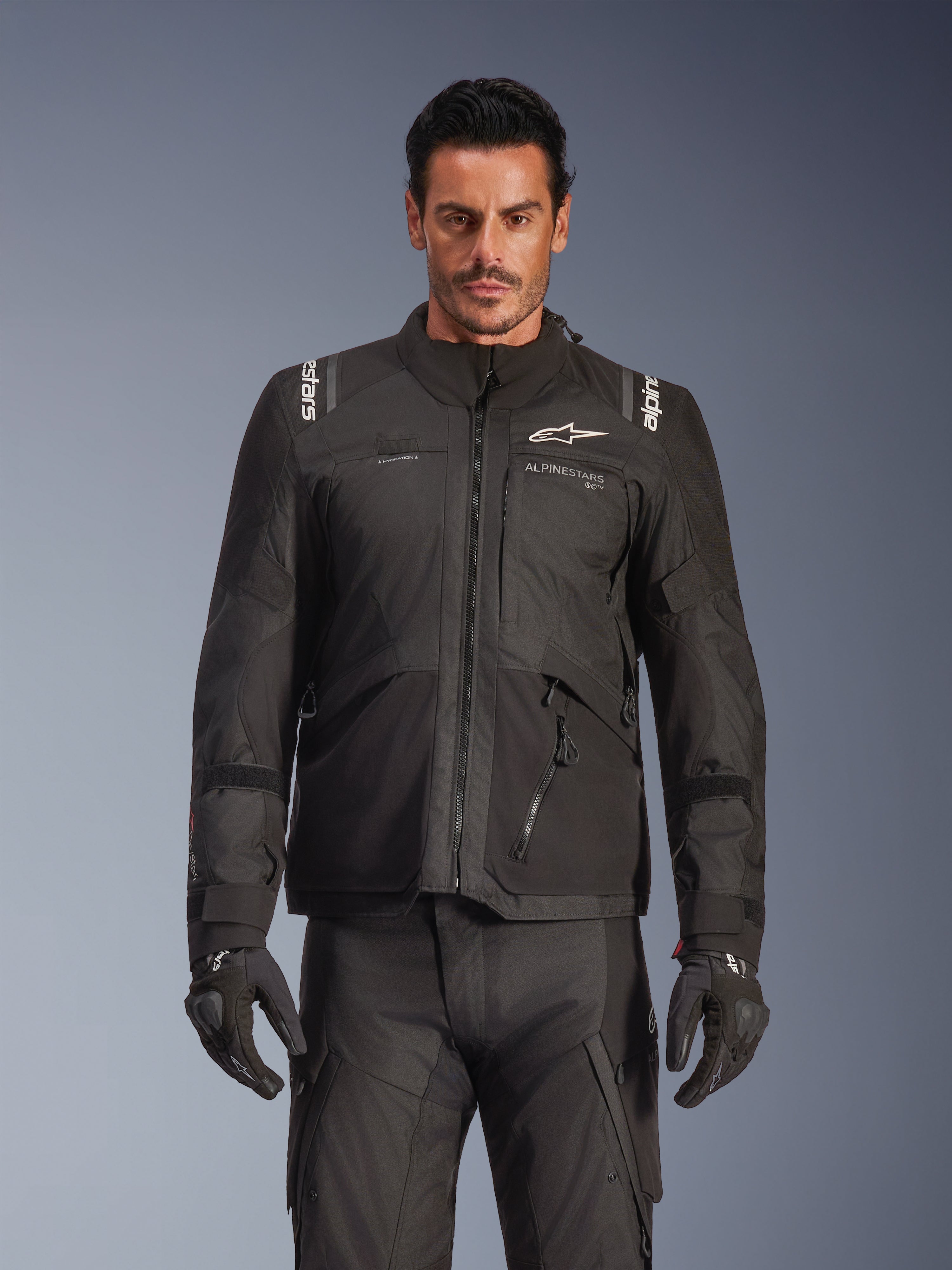 Modelo masculino a usar o Casaco Alpinestars Andes V4 Drystar® em preto, casaco de motociclismo de aventura e turismo, combinado com calças e luvas de condução pretas a condizer, com fechos impermeáveis e logótipo Alpinestars branco no peito e nos ombros, fotografia de estúdio para equipamento de condução em todas as condições meteorológicas.