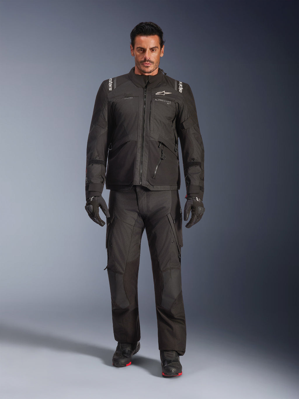 Modelo masculino a usar o Casaco Alpinestars Andes V4 Drystar® em preto, combinado com calças de motociclismo de aventura a condizer, luvas pretas e botas pretas, fotografia de estúdio de corpo inteiro exibindo equipamento técnico de touring para todas as condições climatéricas com a marca Alpinestars.
