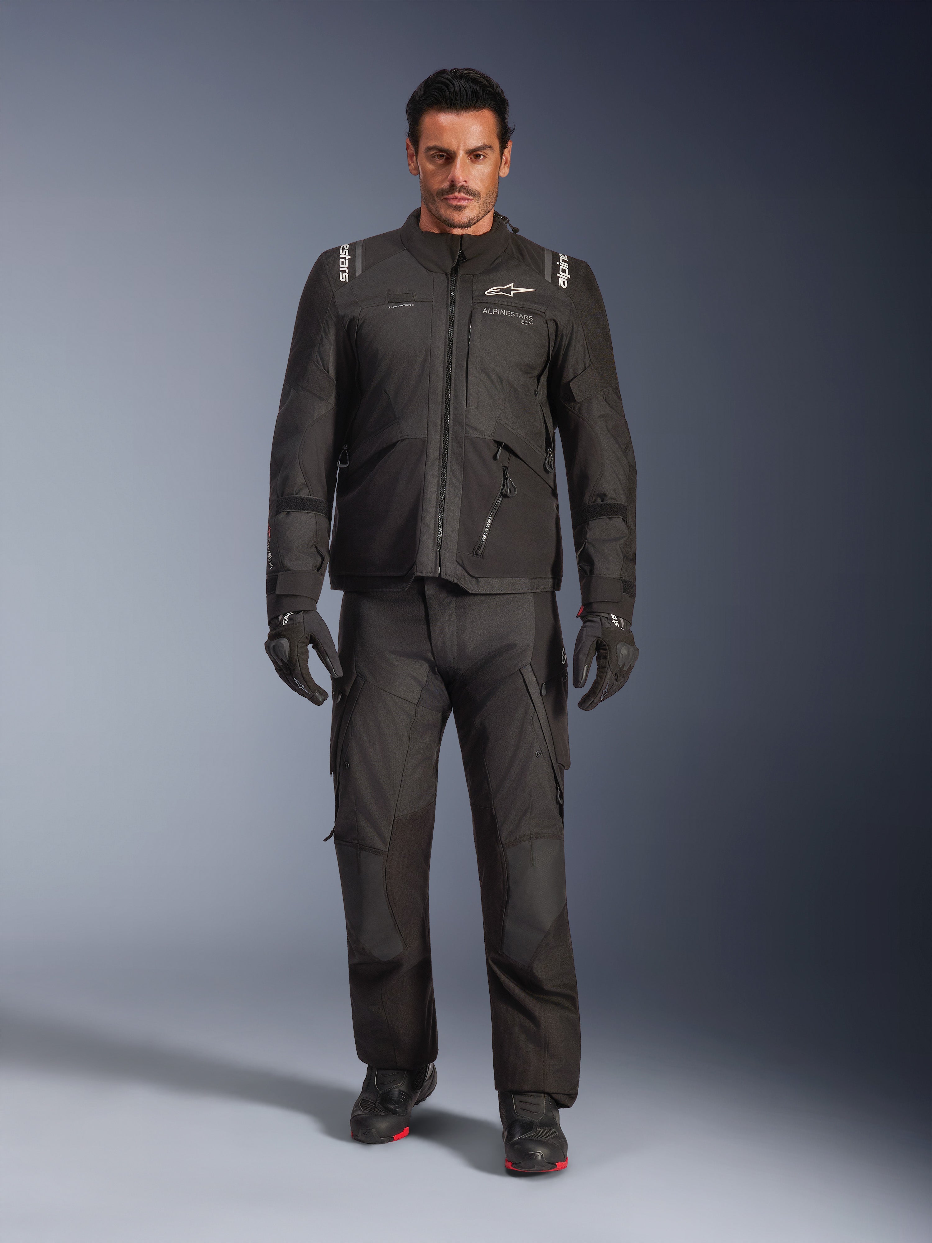 Modelo masculino a usar o Casaco Alpinestars Andes V4 Drystar® em preto, combinado com calças de motociclismo de aventura a condizer, luvas pretas e botas pretas, fotografia de estúdio de corpo inteiro exibindo equipamento técnico de touring para todas as condições climatéricas com a marca Alpinestars.