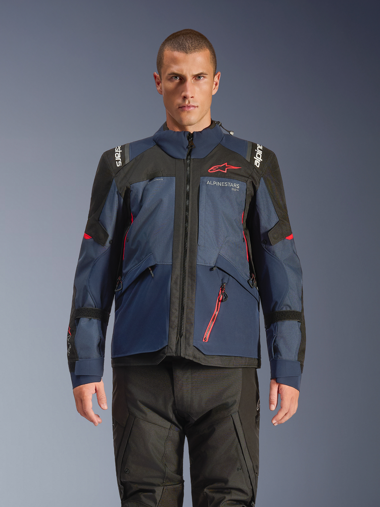 Casaco Alpinestars Andes V4 Drystar®, casaco de motociclismo de aventura, azul Night Navy e Mood Indigo com painéis pretos, vestido por modelo com calças de condução pretas, logótipo Alpinestars vermelho no peito e detalhe de fecho vermelho, construção têxtil durável para turismo em qualquer clima