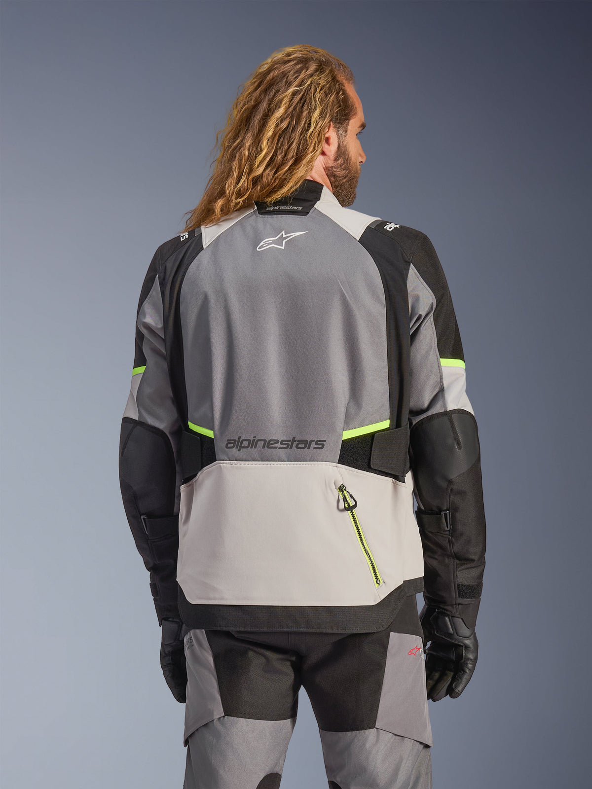 Casaco Alpinestars Andes V4 Drystar®, vista traseira de um casaco de motociclismo de aventura em cinzento escuro, cinzento gelo e preto com detalhes em amarelo néon, com painéis têxteis reforçados e marca Alpinestars, usado com calças a condizer e luvas pretas