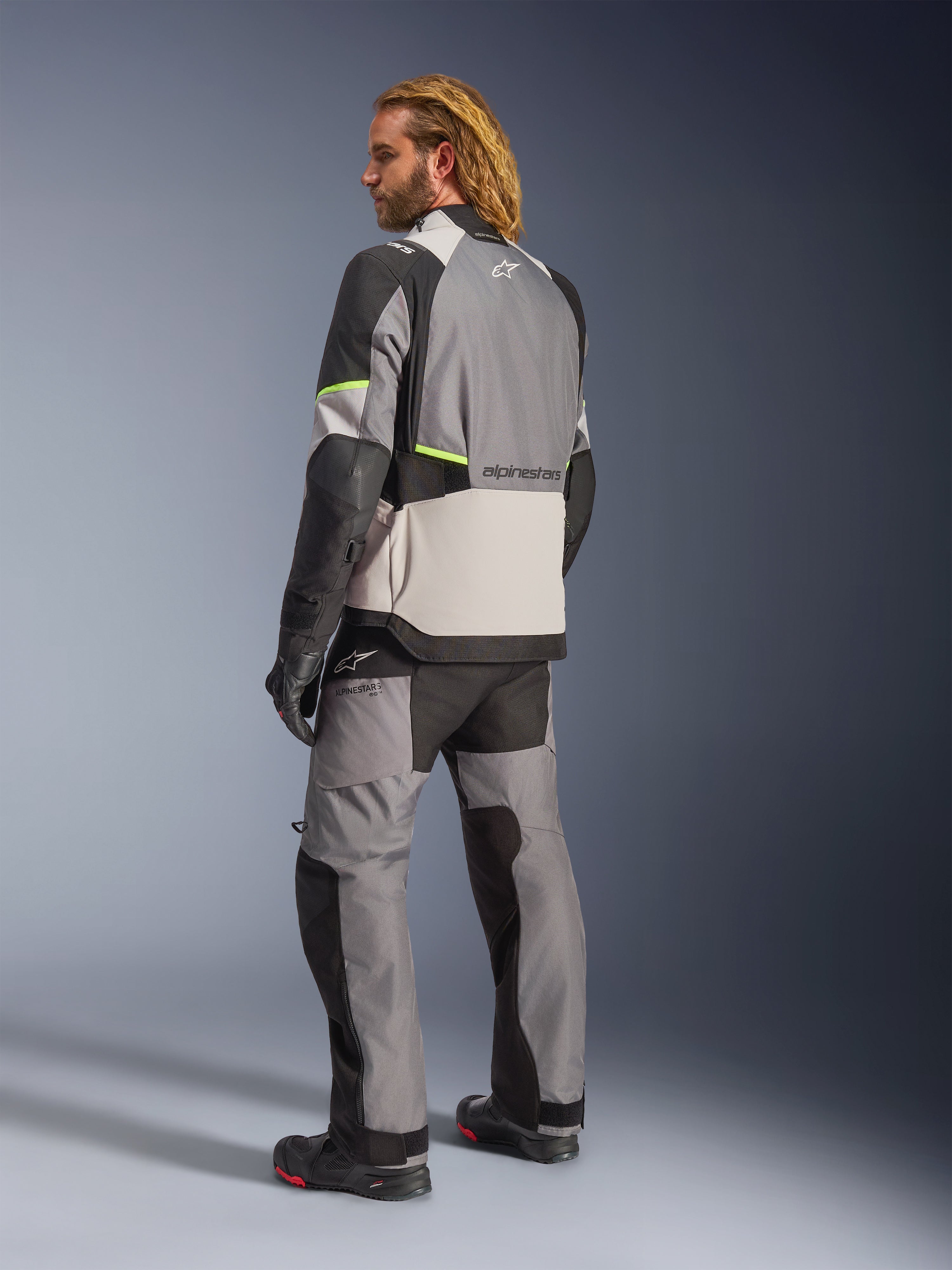 Casaco Alpinestars Andes V4 Drystar®, cinzento escuro, cinzento gelo, preto e amarelo néon, vista traseira de motociclista com calças e botas de touring de aventura a condizer, com painéis têxteis cinzentos reforçados, detalhes em amarelo néon e logótipo Alpinestars, equipamento de motociclismo para todas as condições climatéricas.