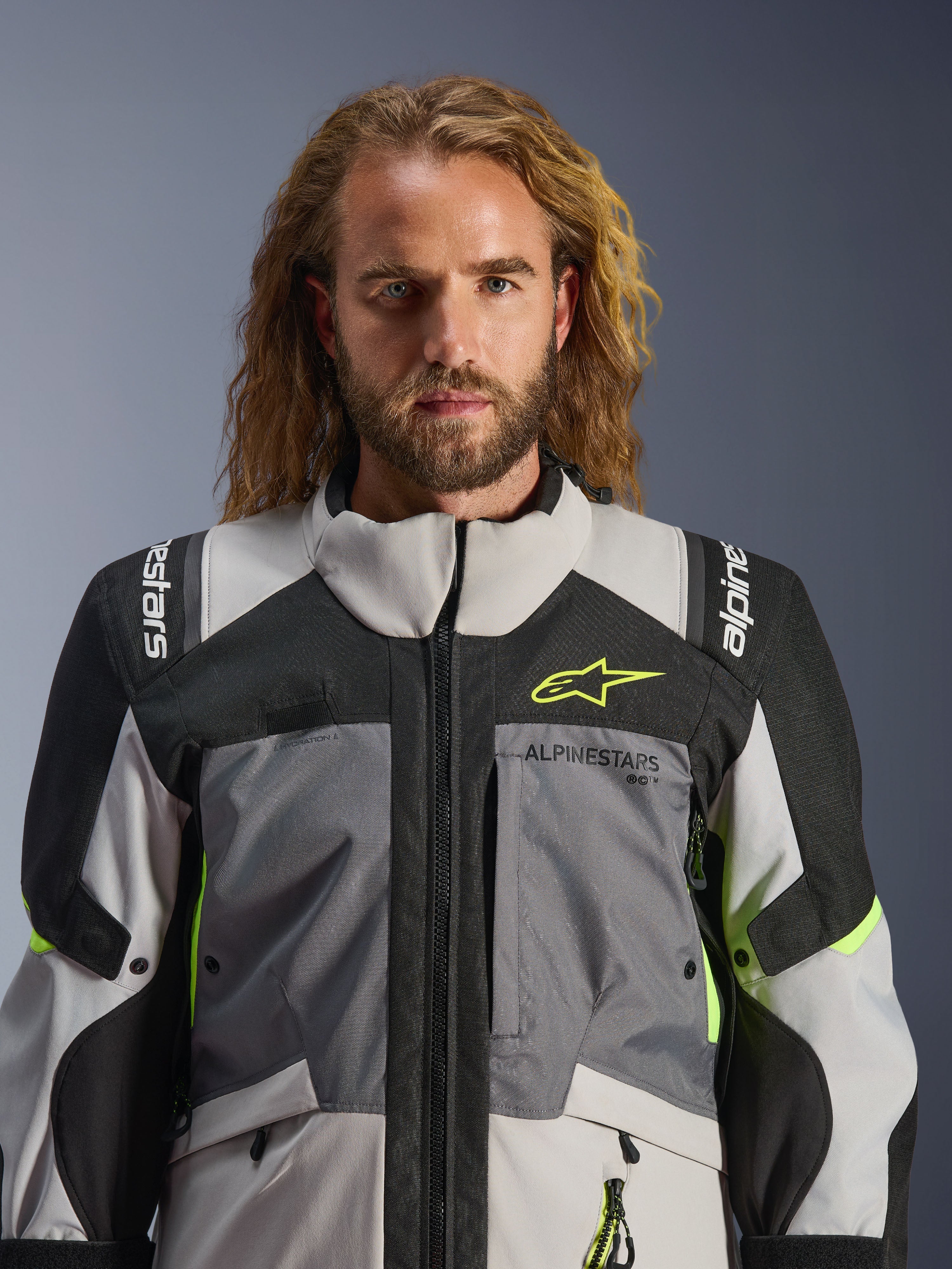 Casaco Alpinestars Andes V4 Drystar®, Cinza Escuro, Cinza Gelo, Preto e Amarelo Fluo, usado por um homem com cabelo comprido e ondulado, apresentando uma construção técnica multi-tecido, logótipo Alpinestars no peito e ombros, e design de gola alta para condução de aventura