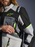 Casaco Alpinestars Andes V4 Drystar®, cinzento escuro, cinzento gelo e preto com pormenores em amarelo flúor, vista aproximada que mostra o bolso de ventilação no peito e a marca Alpinestars, vestido por um motociclista.