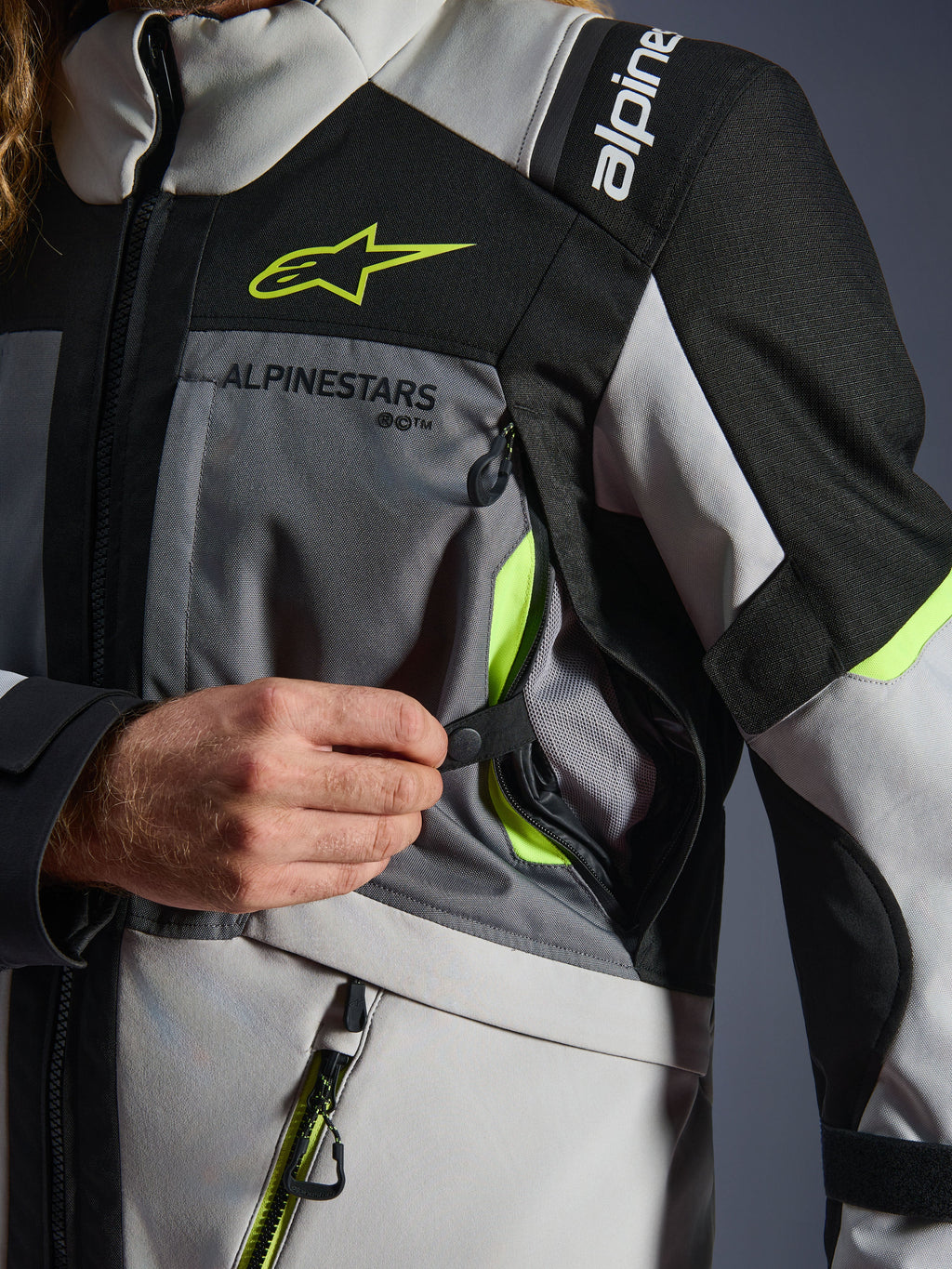 Casaco Alpinestars Andes V4 Drystar®, casaco de motociclismo, cinzento escuro/cinzento gelo/preto/amarelo fluo, pormenor em grande plano que mostra uma mão a abrir a entrada de ventilação no peito com detalhes em amarelo néon e forro em malha, construção têxtil resistente com logótipo Alpinestars.