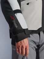 Casaco Alpinestars Andes V4 Drystar®, casaco de motociclismo de aventura, painéis em cinzento gelo e preto, grande plano da manga com o logótipo Drystar® vermelho e correias ajustáveis, design têxtil funcional para condução em todas as condições climáticas