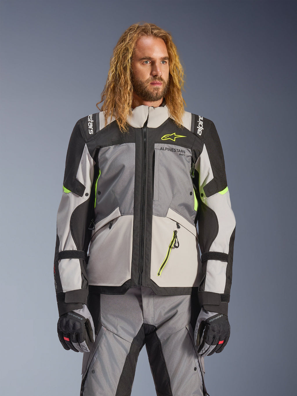 Casaco Alpinestars Andes V4 Drystar, casaco de moto para turismo de aventura, cinzento escuro e cinzento gelo com detalhes em amarelo néon, vestido por um modelo com calças e luvas a condizer, design têxtil multibolsos para condução em todas as condições climáticas