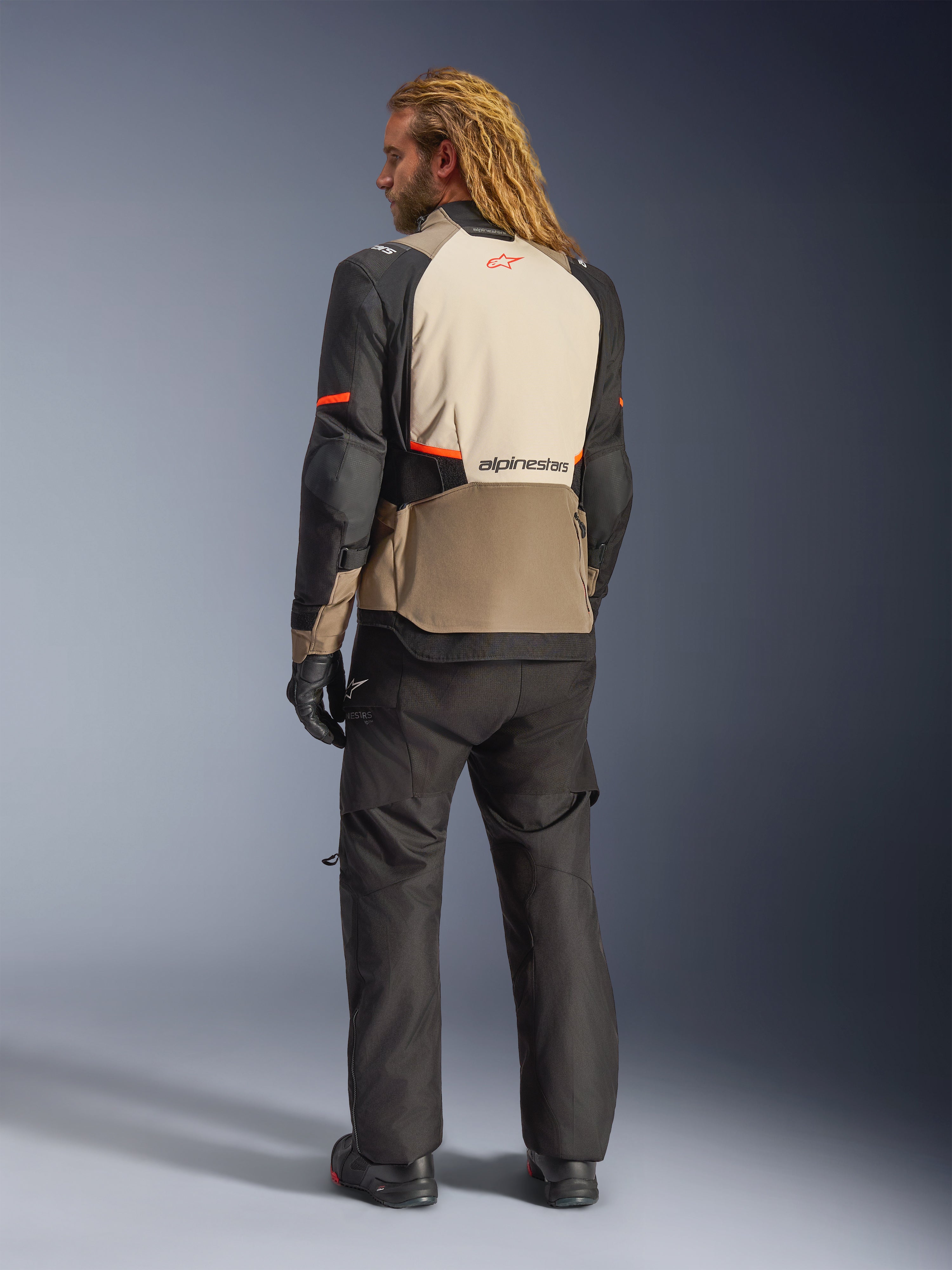 Casaco Alpinestars Andes V4 Drystar®, Walnut Mastic Black, vista traseira num modelo masculino, com painéis têxteis reforçados em bege e preto com detalhes laranja, logótipo Alpinestars nas costas, usado com calças de aventura e luvas pretas a condizer.