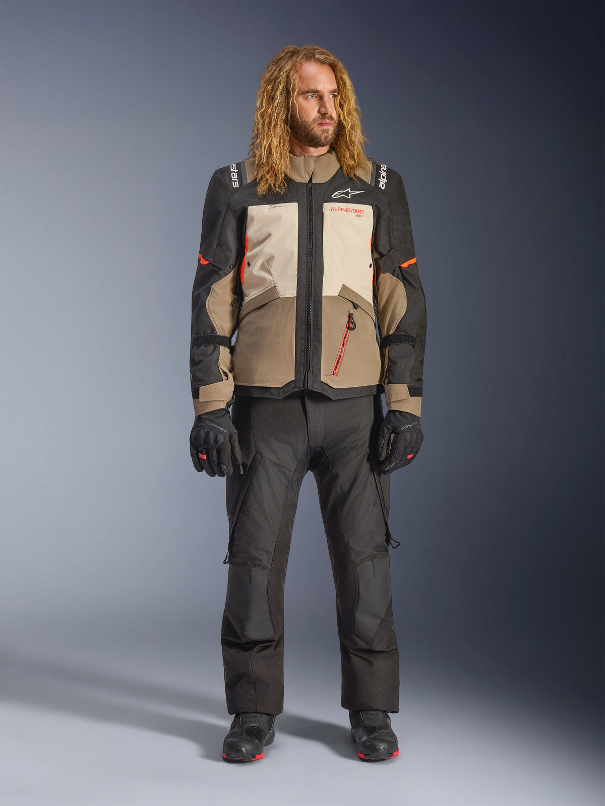 Casaco Alpinestars Andes V4 Drystar®, Walnut Mastic Black, usado com calças e luvas de motociclismo pretas, com um design de aventura multi-painel com detalhes laranja e logótipos Alpinestars, equipamento têxtil técnico para condução em todas as condições climatéricas