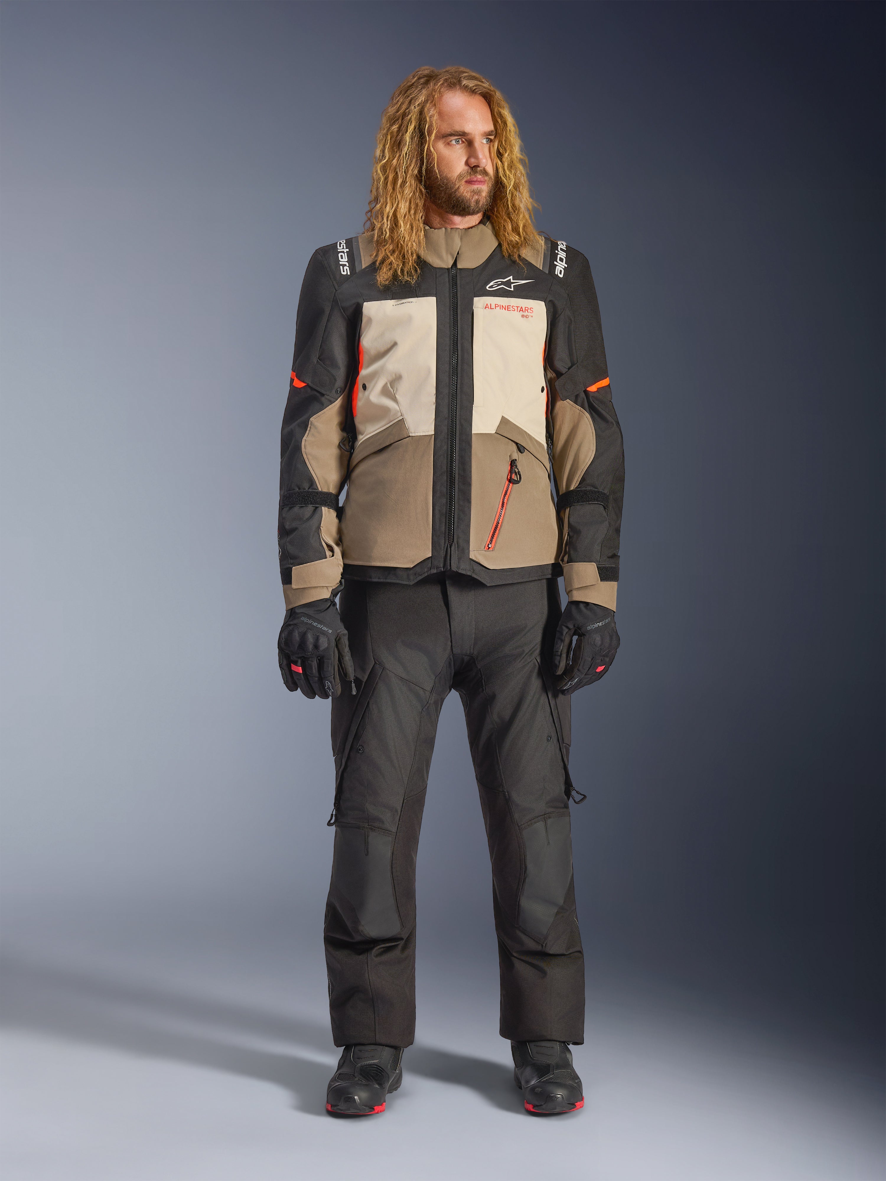 Casaco Alpinestars Andes V4 Drystar®, Walnut Mastic Black, usado com calças e luvas de motociclismo pretas, com um design de aventura multi-painel com detalhes laranja e logótipos Alpinestars, equipamento têxtil técnico para condução em todas as condições climatéricas