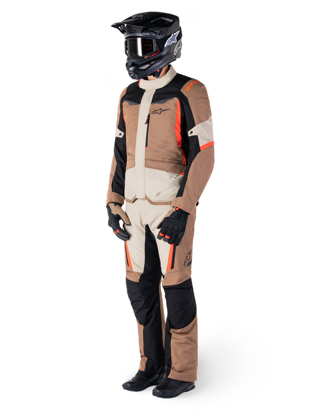 Casaco Impermeável Alpinestars ST-1, cáqui escuro, areia, preto e vermelho fluorescente, usado por um motociclista com equipamento completo de turismo de aventura com calças a condizer, capacete preto, óculos, luvas e botas, conjunto técnico de motociclismo para todas as condições climatéricas