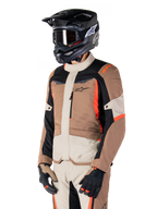 Casaco Alpinestars ST-1 Waterproof, casaco de touring de aventura, Dark Khaki Sand Black e Red Fluo, usado por um piloto com capacete de motocross preto e óculos de proteção, com design de vários painéis, bolsos cargo e logótipo Alpinestars no peito
