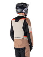 Casaco Impermeável Alpinestars ST-1, casaco de turismo de aventura, cáqui escuro, areia, preto e vermelho fluorescente, vista traseira com painéis têxteis em blocos de cor, detalhes em vermelho fluo e marca Alpinestars na zona inferior das costas, usado por um motociclista com capacete preto