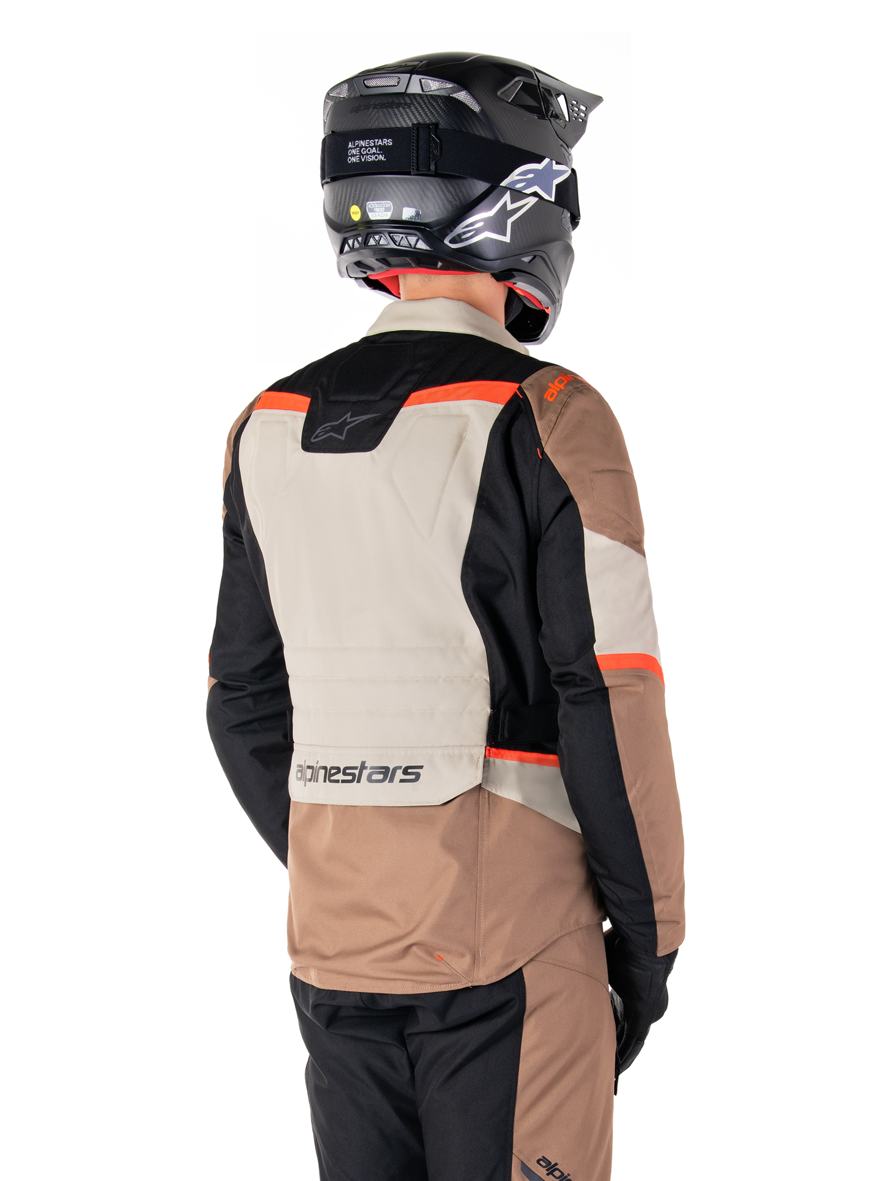 Casaco Impermeável Alpinestars ST-1, casaco de turismo de aventura, cáqui escuro, areia, preto e vermelho fluorescente, vista traseira com painéis têxteis em blocos de cor, detalhes em vermelho fluo e marca Alpinestars na zona inferior das costas, usado por um motociclista com capacete preto