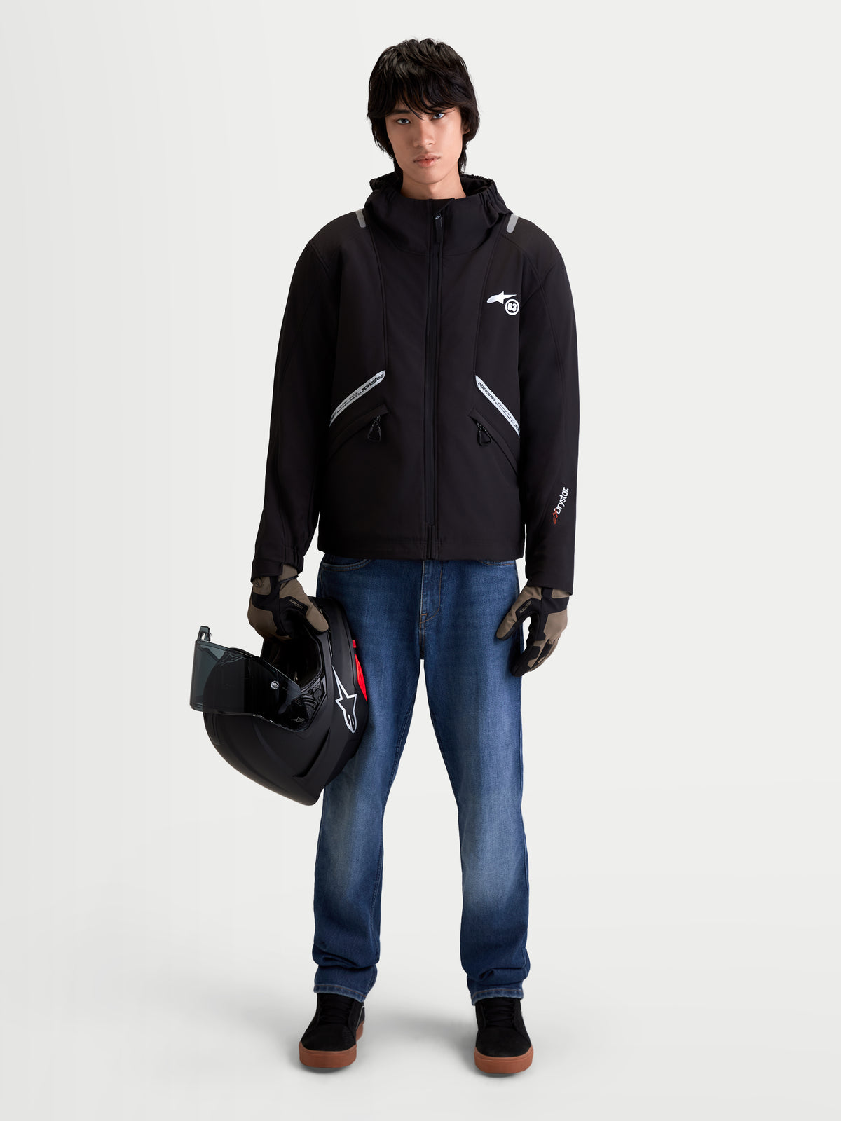 Alpinestars C 5 Drystar casaco de moto urbano para homem, preto, vista frontal num modelo a usar calças de ganga e luvas, segurando um capacete, destacando a construção em softshell impermeável com capuz integrado