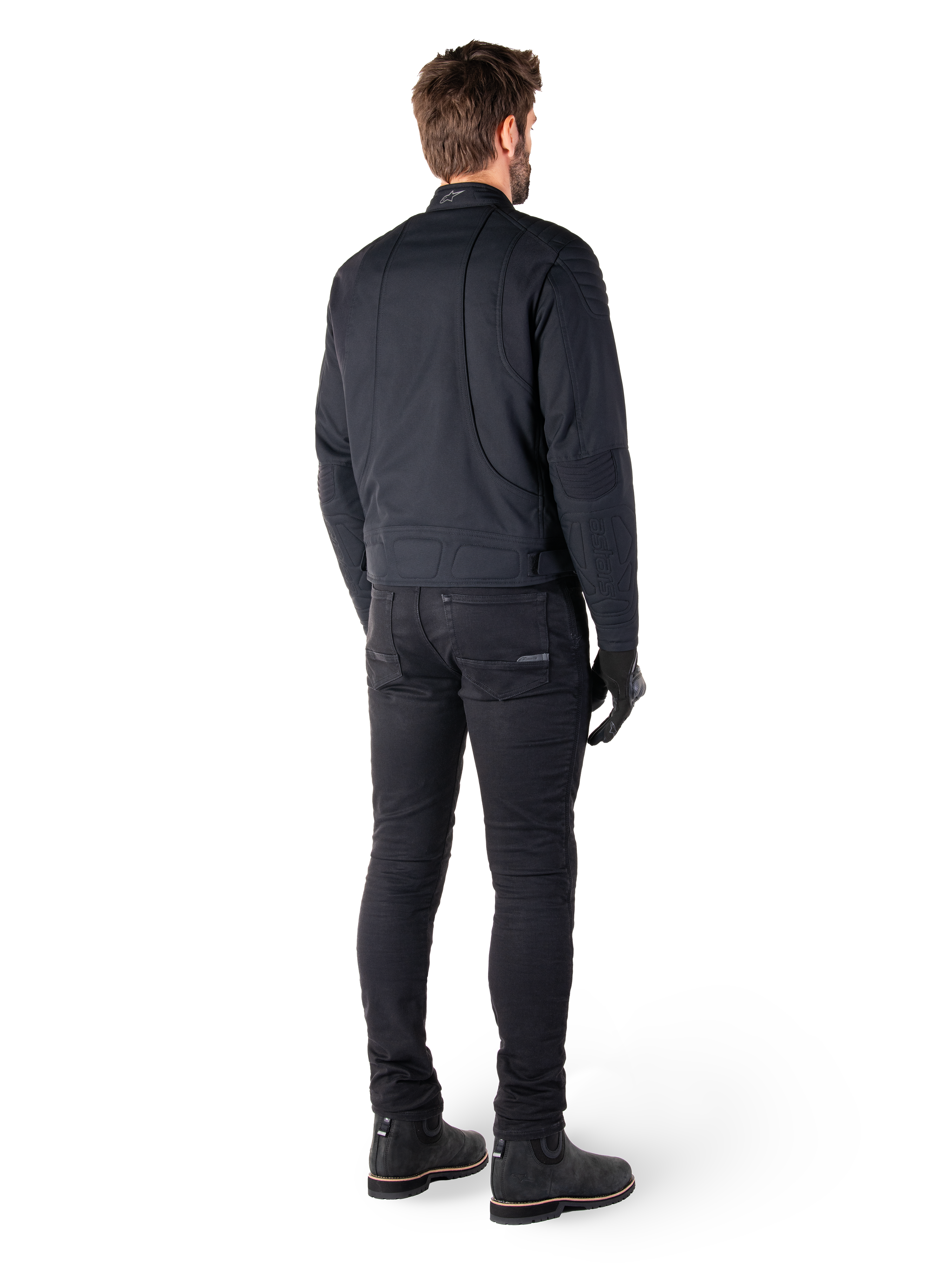 Vista traseira de um motociclista a usar o Casaco Alpinestars Clayton WR em preto, com painéis acolchoados nos cotovelos e ombros, costuras curvas ergonómicas e detalhes refletores na gola, combinado com calças e botas de motociclismo pretas para um visual urbano.