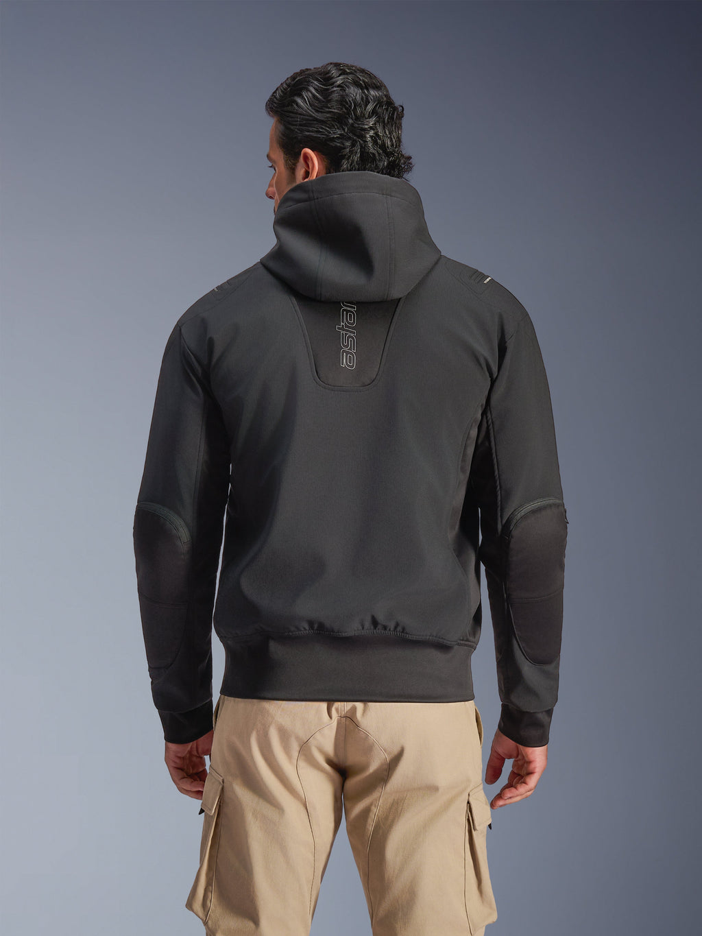 Alpinestars Moblast Waterproof Jacket, preto, vista traseira de casaco de motociclismo urbano com capuz, apresentando logótipo 'astars' vertical nas costas, cintura e punhos elásticos, cotovelos reforçados e design moderno e tecnológico, usado com calças cargo bege