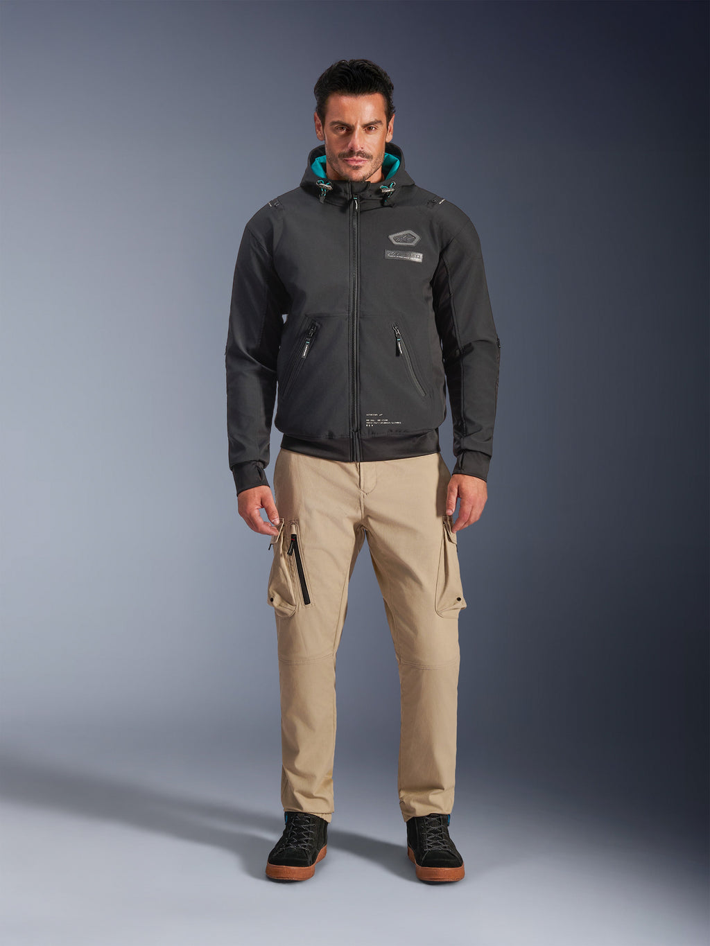 Casaco Impermeável Alpinestars Moblast, hoodie de motociclismo urbano preto com forro interior do capuz em azul-petróleo, usado com calças cargo bege e botas de cano alto pretas, vestuário de condução contemporâneo para todas as estações