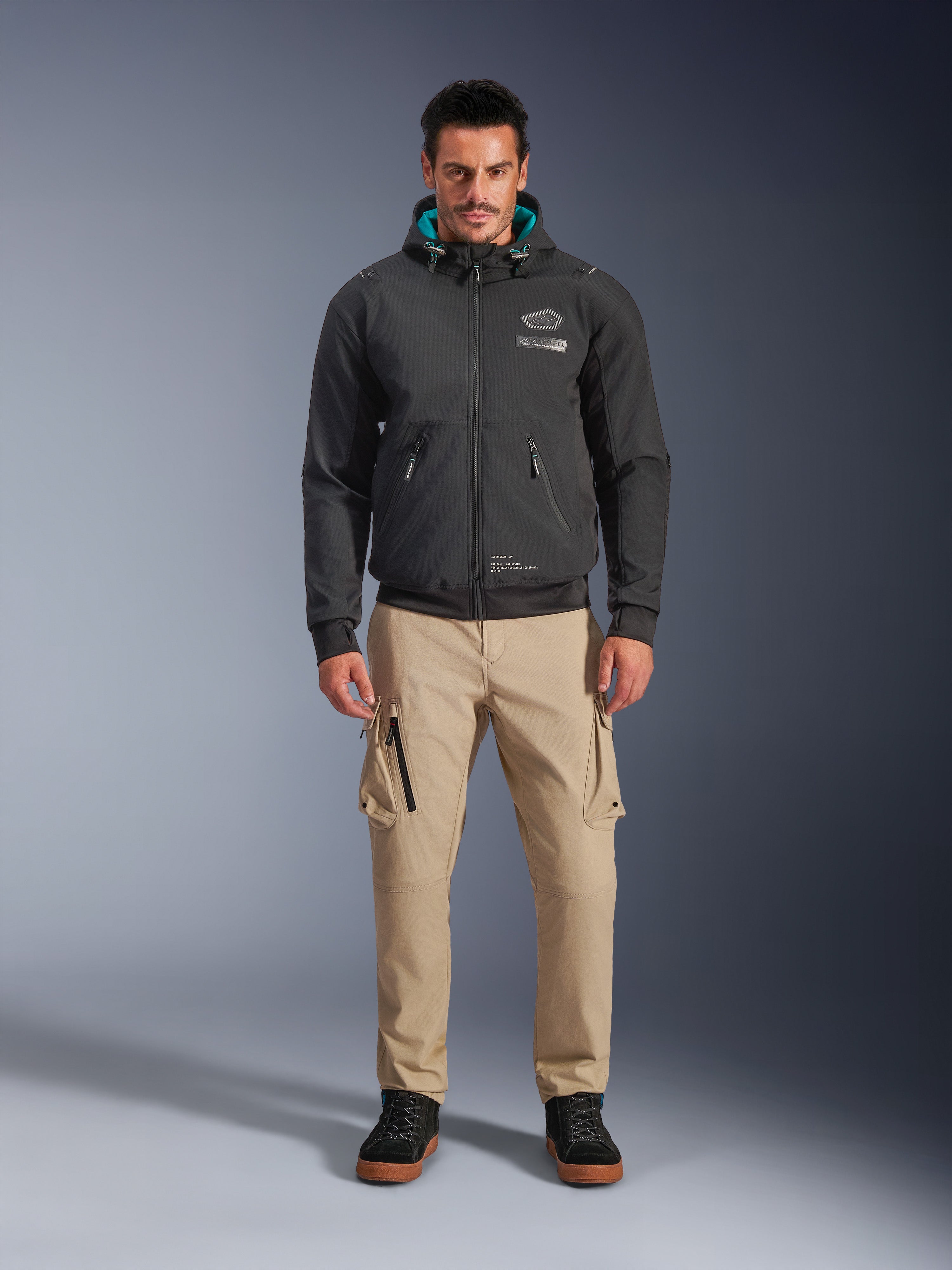 Casaco Impermeável Alpinestars Moblast, hoodie de motociclismo urbano preto com forro interior do capuz em azul-petróleo, usado com calças cargo bege e botas de cano alto pretas, vestuário de condução contemporâneo para todas as estações