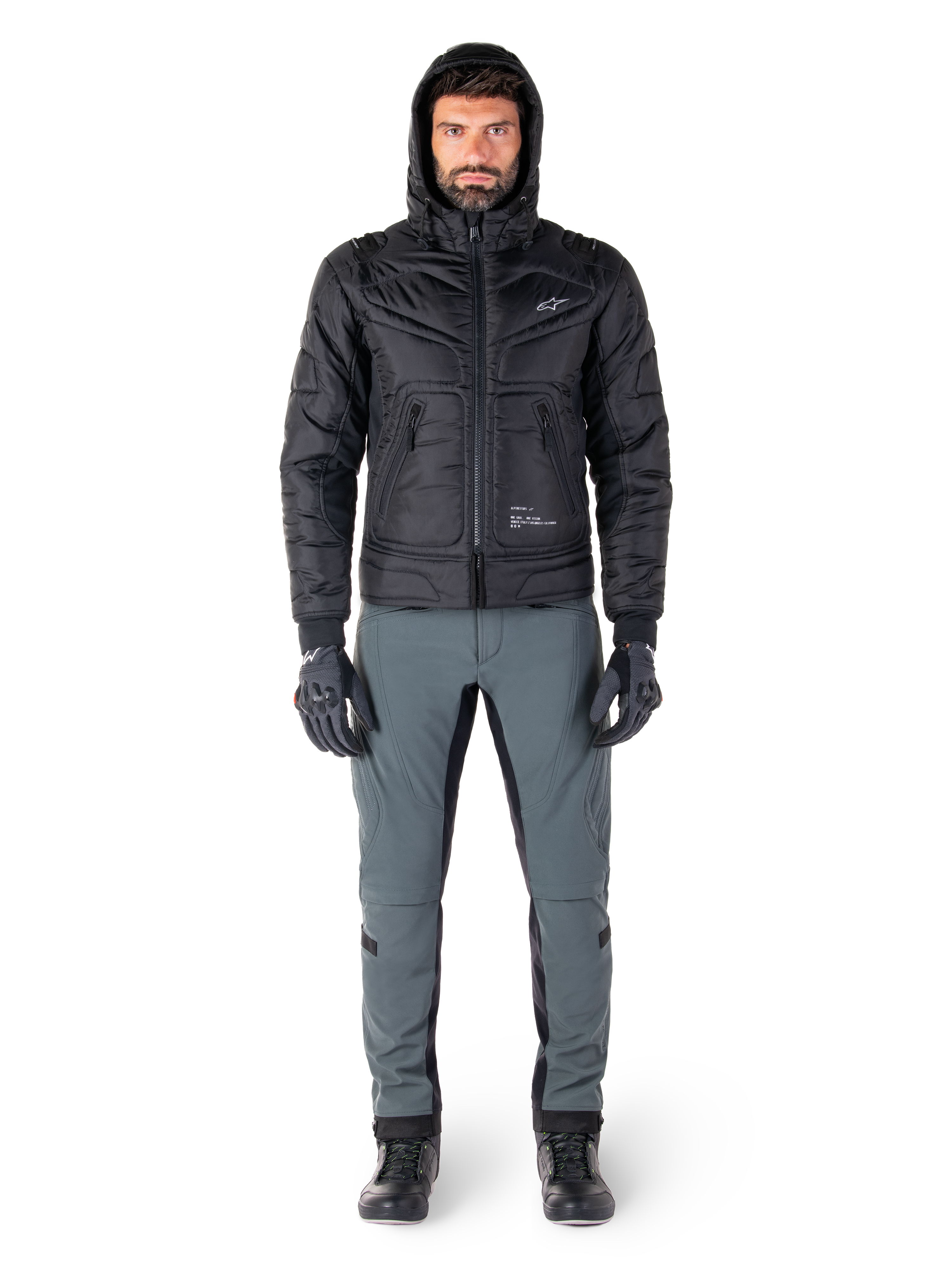 Alpinestars Mohobbs Waterproof Jacket in black, male model wearing quilted padded bomber jacket with hood, paired with grey riding pants, black gloves, and black boots, urban contemporary motorcycle gear.