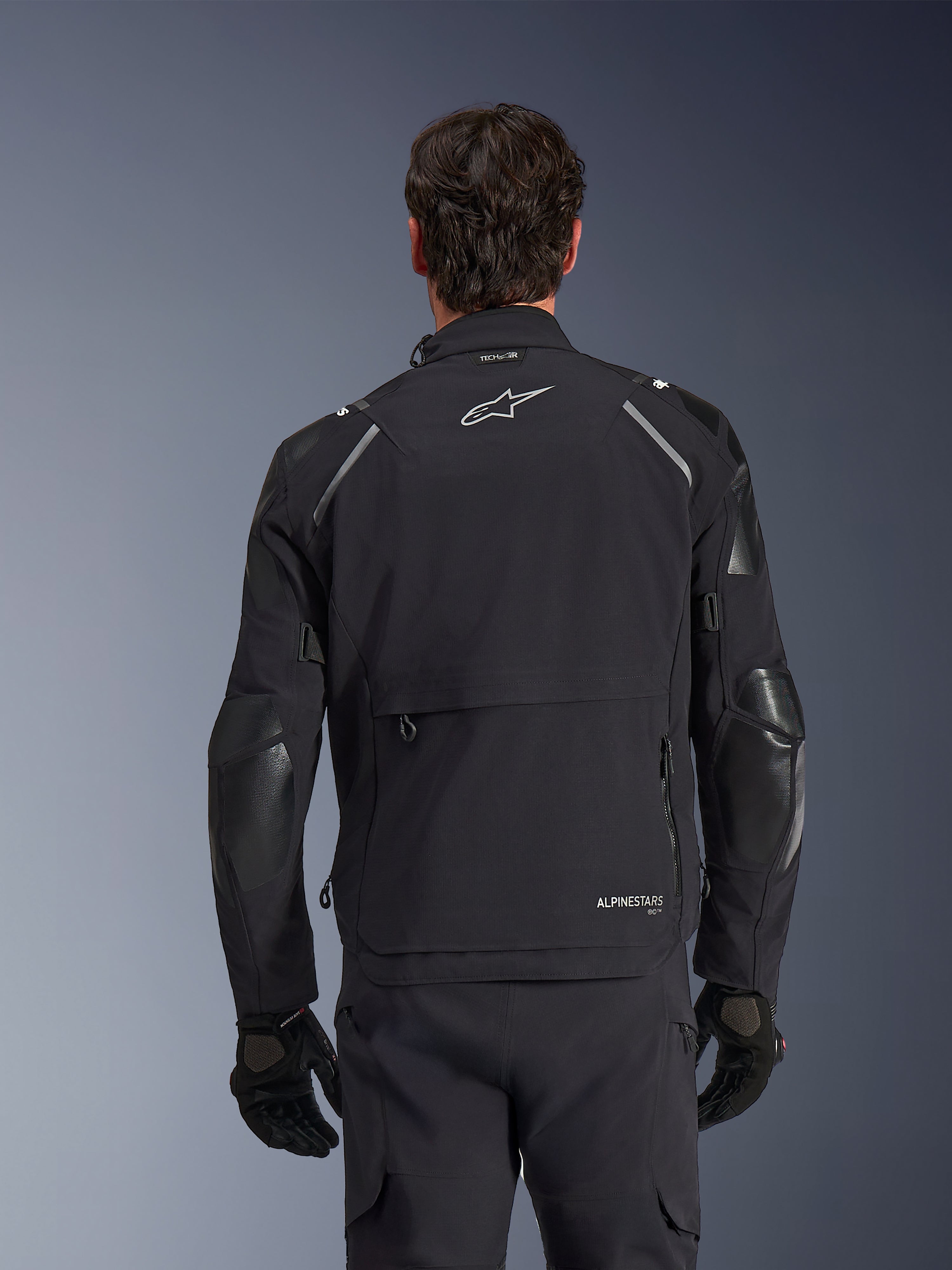 Casaco Alpinestars Andes Pro Drystar® XF, preto e cinzento escuro, vista traseira num modelo masculino, com um compartimento de armazenamento traseiro grande, detalhes refletores nos ombros e logótipo da marca, combinado com luvas e calças pretas para motociclismo de aventura.